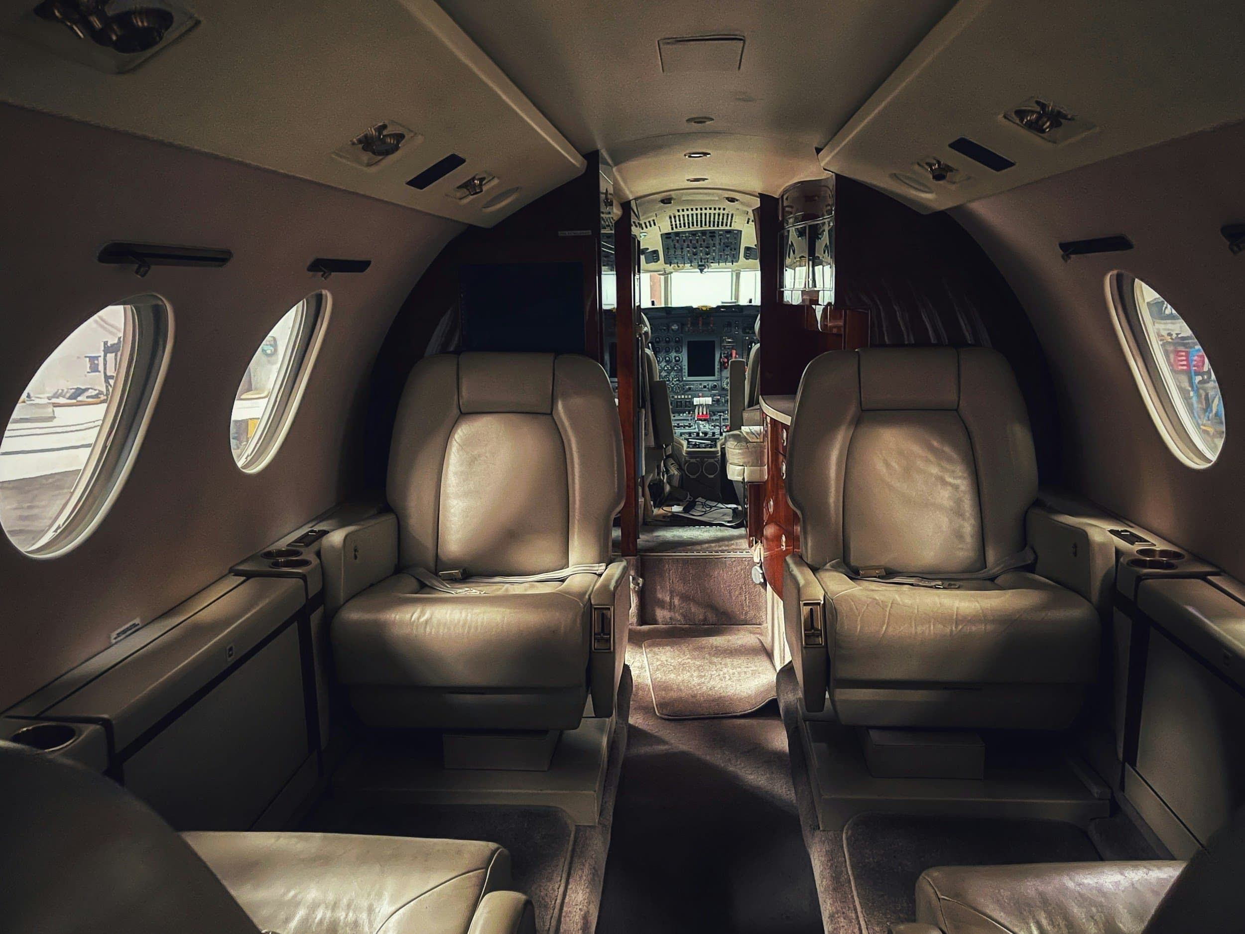Interior of a private jet with beige leather seats, multiple windows, and a cockpit visible in the background.